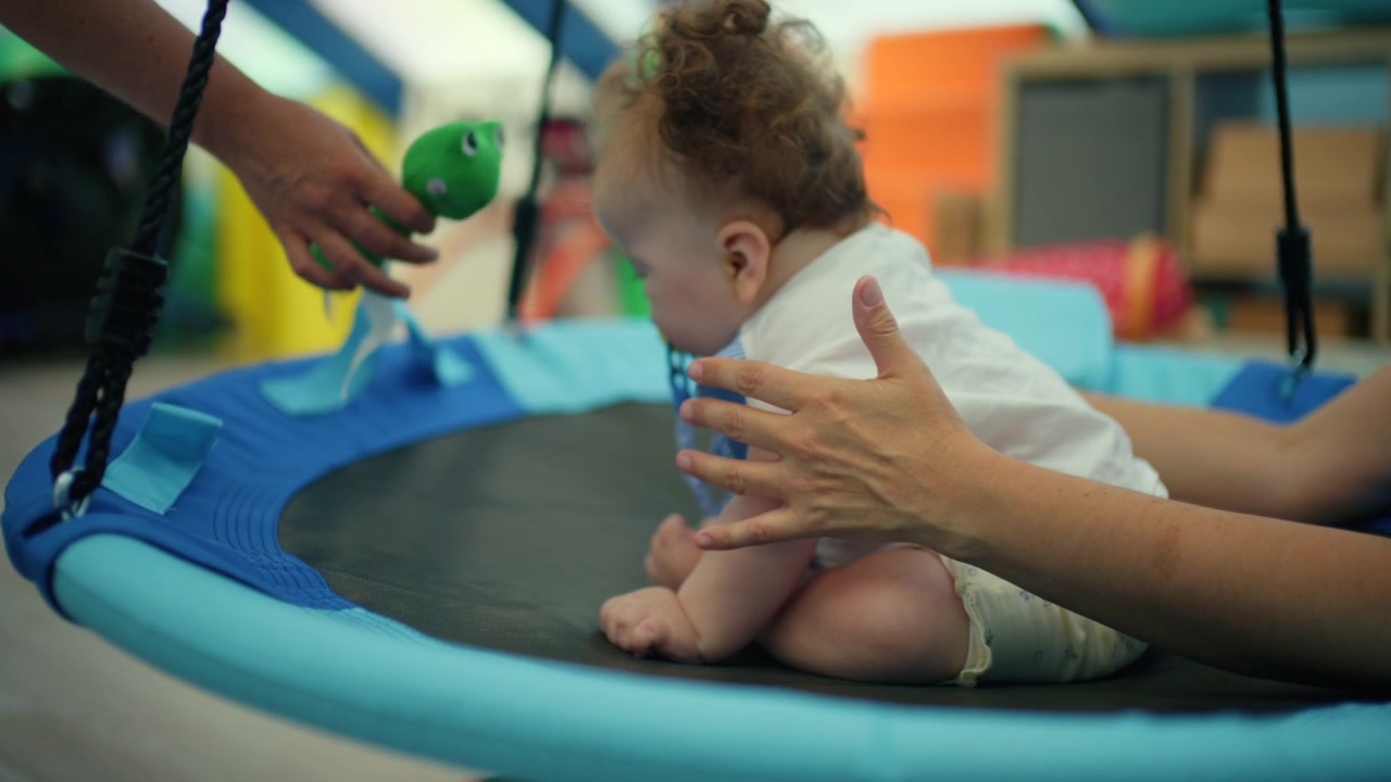 Bebê sentado em um balanço azul, aprendendo e brincando durante uma sessão de fisioterapia com um instrutor