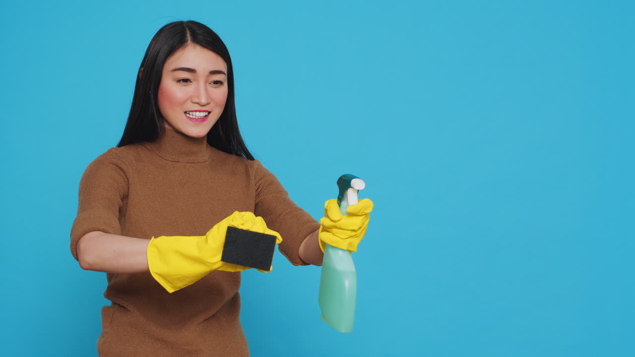 Happy asian maid wearing protective gloves while cleaning house using sponge and detergent spray