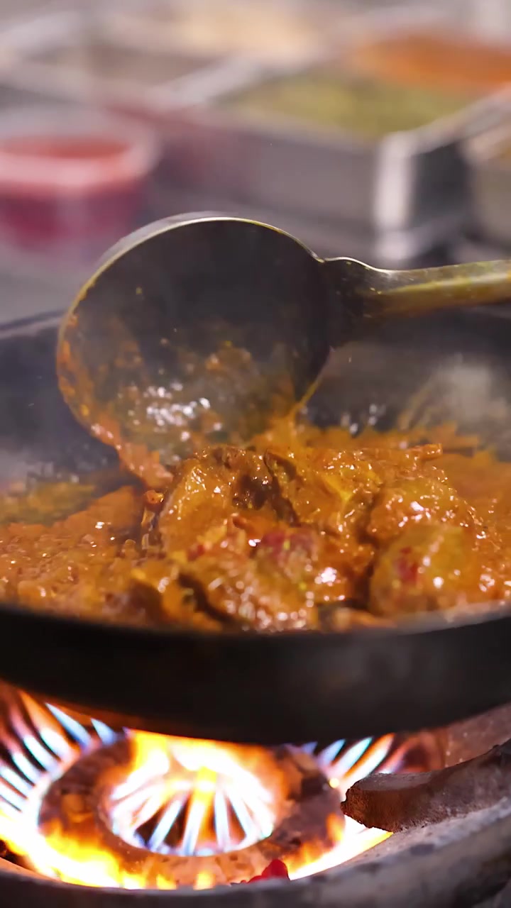 Primo piano di uno chef che mescola un curry di carne che sobbolle in una cucina professionale su una fiamma a gas alta.