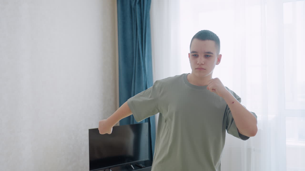 Focused teen boy performs arm flexibility workout at home near window with sheer curtains, wearing loose shirt, he moves arms deliberately, maintaining posture and balance