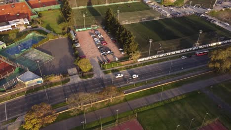 Toma-Aérea-De-Dos-Autos-Blancos-Conduciendo-En-La-Avenida-Al-Lado-Del-Campo-De-Fútbol-De-Buenos-Aires-Durante-La-Puesta-De-Sol