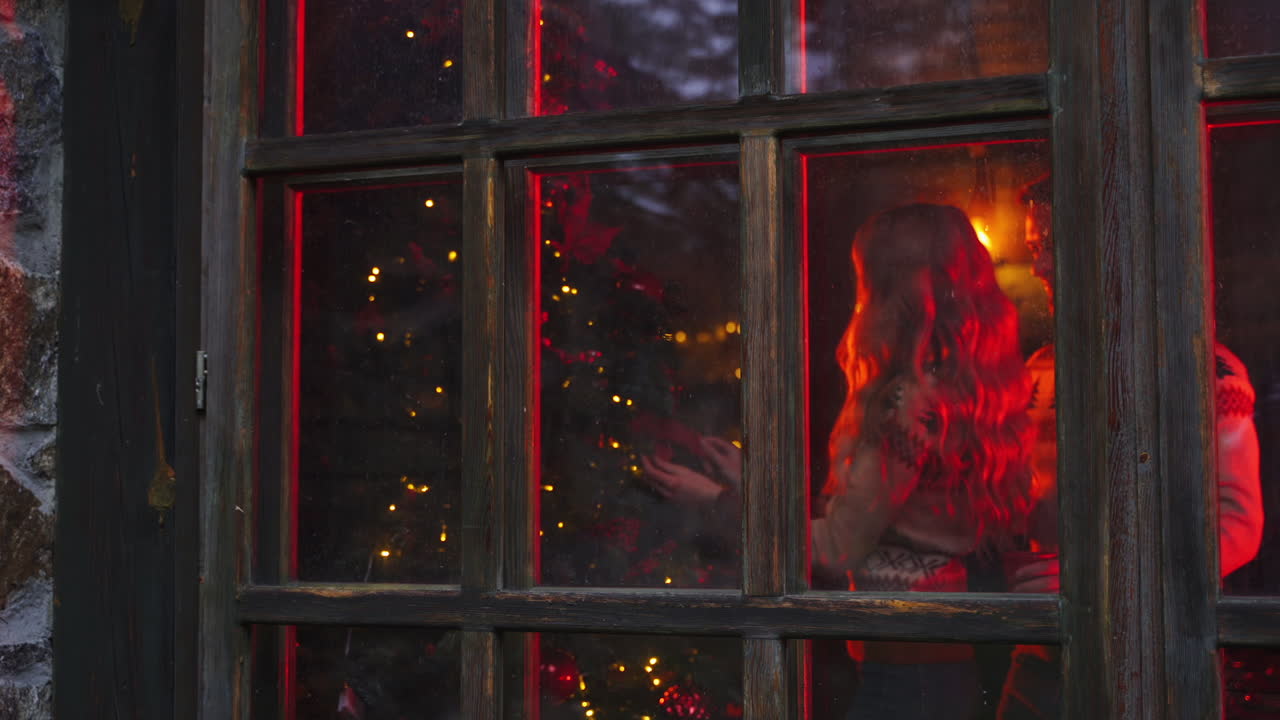 Comfortable atmosphere inside the house. Young family prepares for New Year. Woman decorates Christmas tree near the window. Husband brings hot tea. View from outdoors.