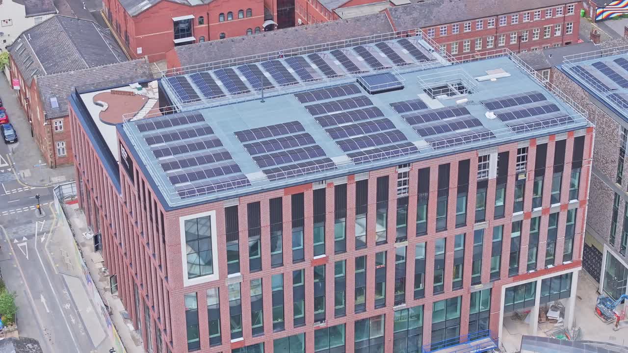 An aerial shot of a building with solar panels covering its rooftop. A construction site with cranes and workers can be seen nearby. The building is located in a city, surrounded by other buildings