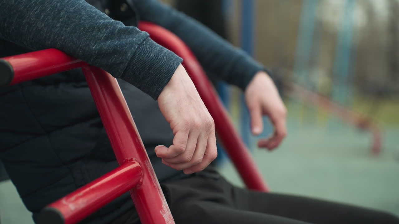 Close-up of a person's hands resting on a workout equipment as he shakes his leg, with a blurred view of a boy playing football and blue poles in the background