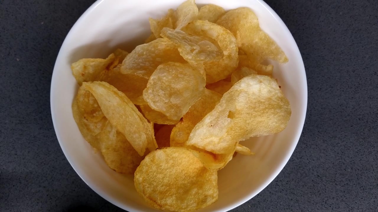 Hand pours potato chips into a white bowl and picks one up on a dark countertop