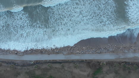 Top-drone-shot-captures-road-leading-along-the-coast-line-as-white-foamy-waves-wash-ashore