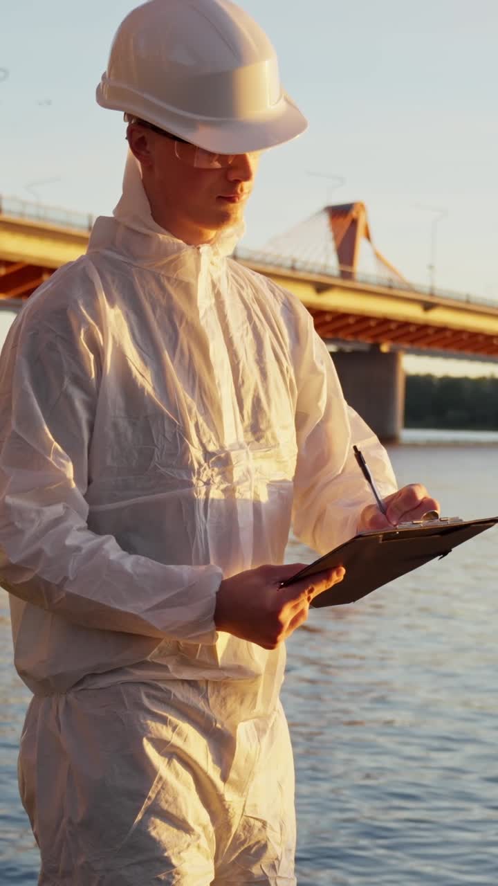 Inspector standing near industrial structure assessing surroundings for environmental impact holding clipboard taking notes at sunset, vertical