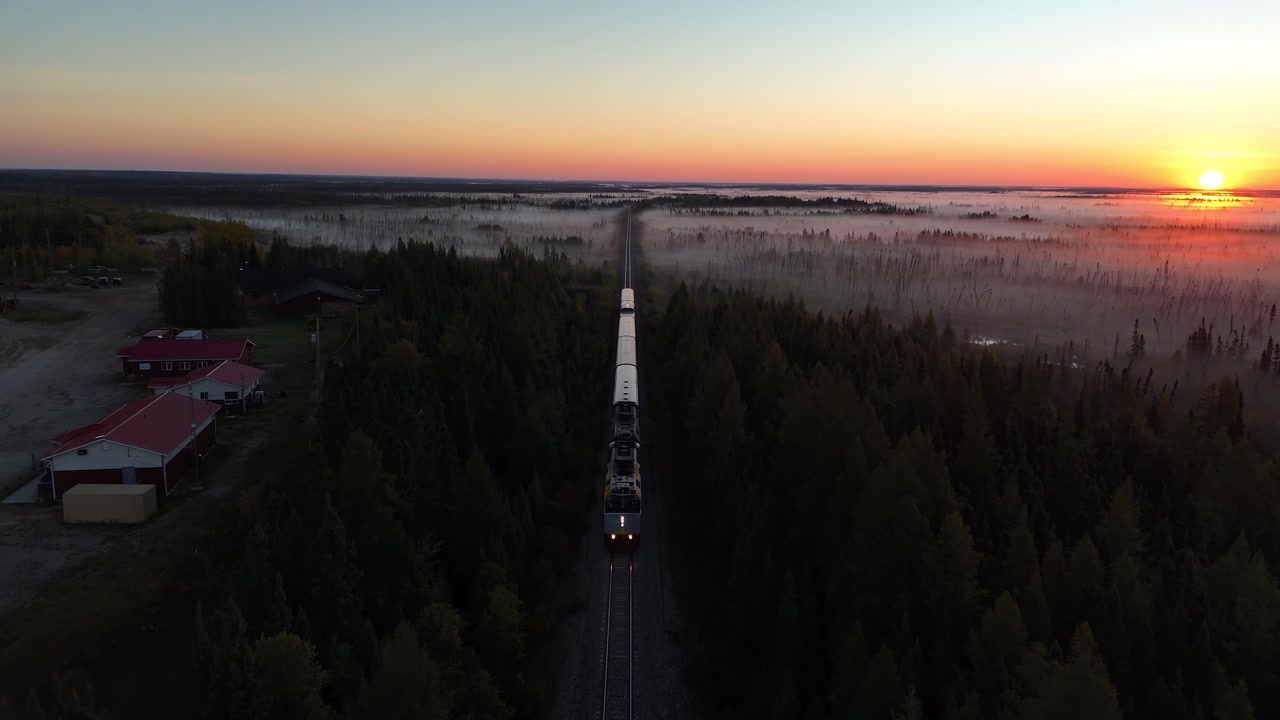 Aerial drone shot flying overhead as Via Rail train from Churchill approaches War Lake Ilford station in northern Manitoba. Sunrise light with misty boreal forest and nearby community buildings.
