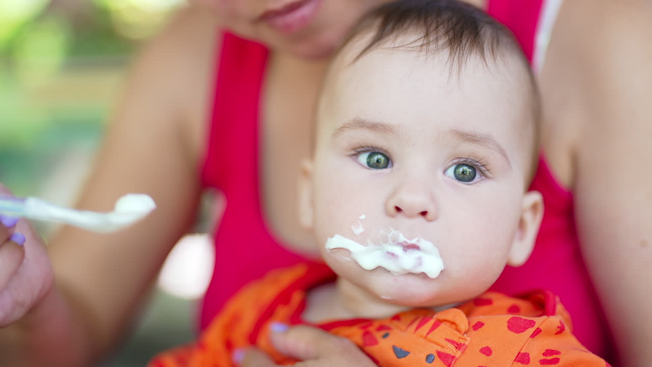 Beautiful baby murky from food he is being fed with. Mom gives a spoon to her child but he turns the head away. Close up.