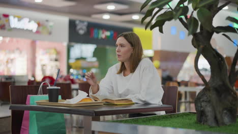 primer plano de una mujer comiendo un bocadillo mientras lee un libro en un restaurante moderno, fondo vibrante con iluminación suave, bebida en la mesa y bolsas de compras a la vista