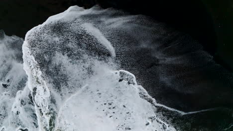 Side-angle,-birds-eye-view-aerial-shot-of-shite-tide-crashing-on-contrasting-black-sand-beach-in-Bali