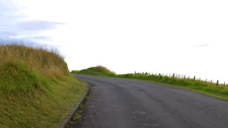 Walking-through-asphalted-road-to-skyline-in-green-grass-hills-wild-landscape-POV-point-of-view