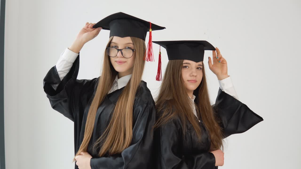 Two young happy graduate women stand shoulder to shoulder correct their hats with their hands. Close up view on white background