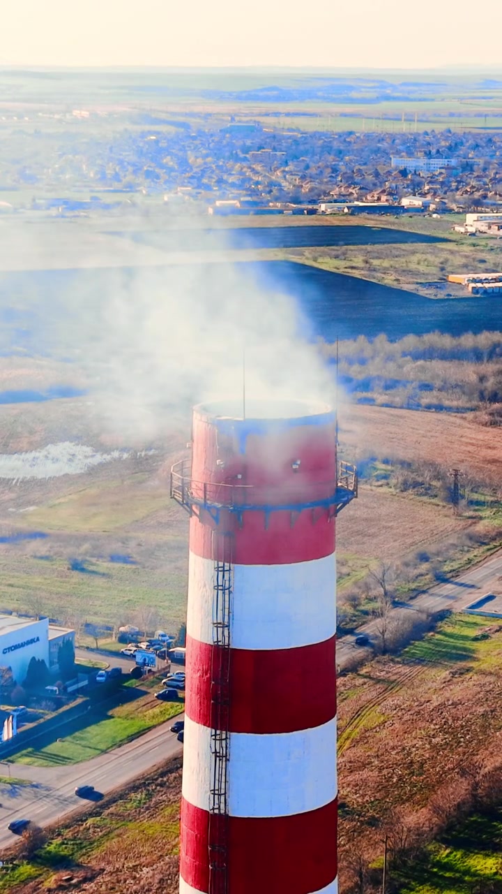 Vista aerea di camino a strisce con fumo su una città bulgara. Alta torre industriale si erge sopra i tetti, la strada e un campo verde sotto un cielo velato. Video verticale