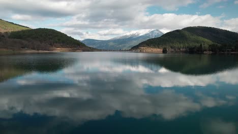 Nubes-Reflejadas-En-Las-Aguas-Del-Lago-Artificial-Doxa-Entre-Colinas-Verdes-En-Grecia-Drones-Aéreos-Volando-Cerca-De-La-Superficie,-Escenario-Pintoresco