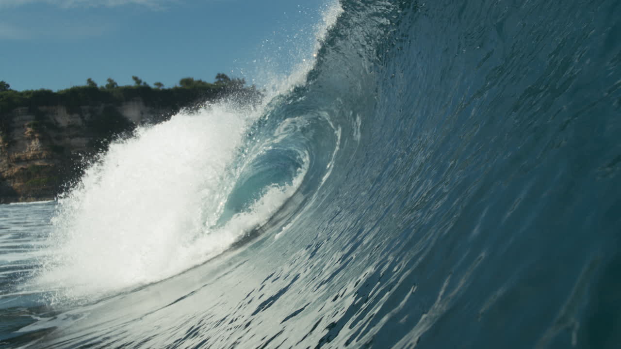 Rolling wave breaks in sunlight with ripples trailing behind in open water, glistening water