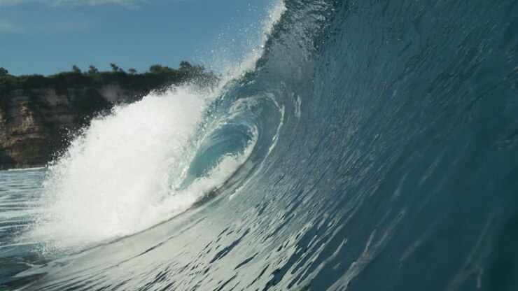 Rolling wave breaks in sunlight with ripples trailing behind in open water, glistening water