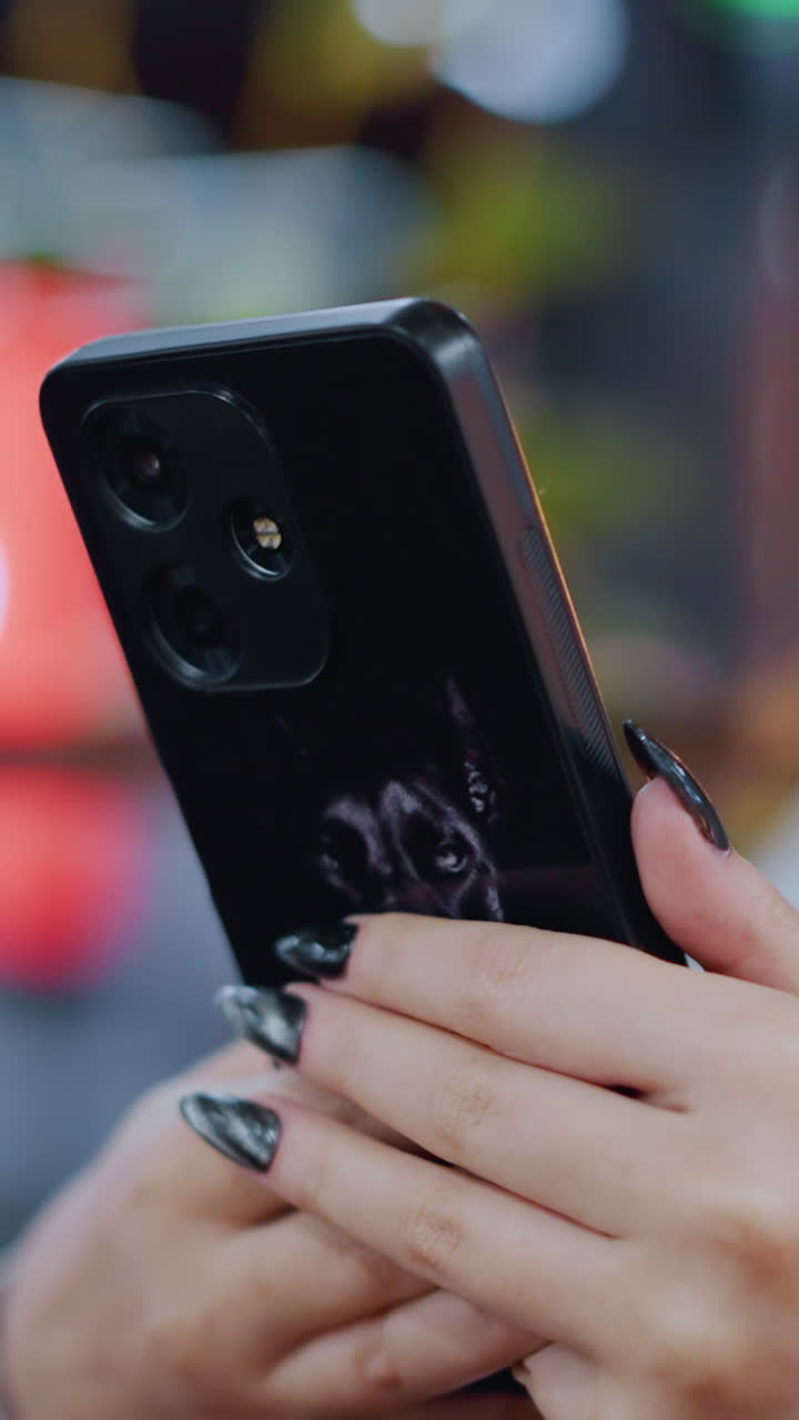 primer plano de uñas pintadas de color negro sosteniendo un iphone con pegatina de burro mientras el propietario lo opera con un fondo borroso, centrarse en la funda personalizada del teléfono y uñas elegantes en un entorno casual
