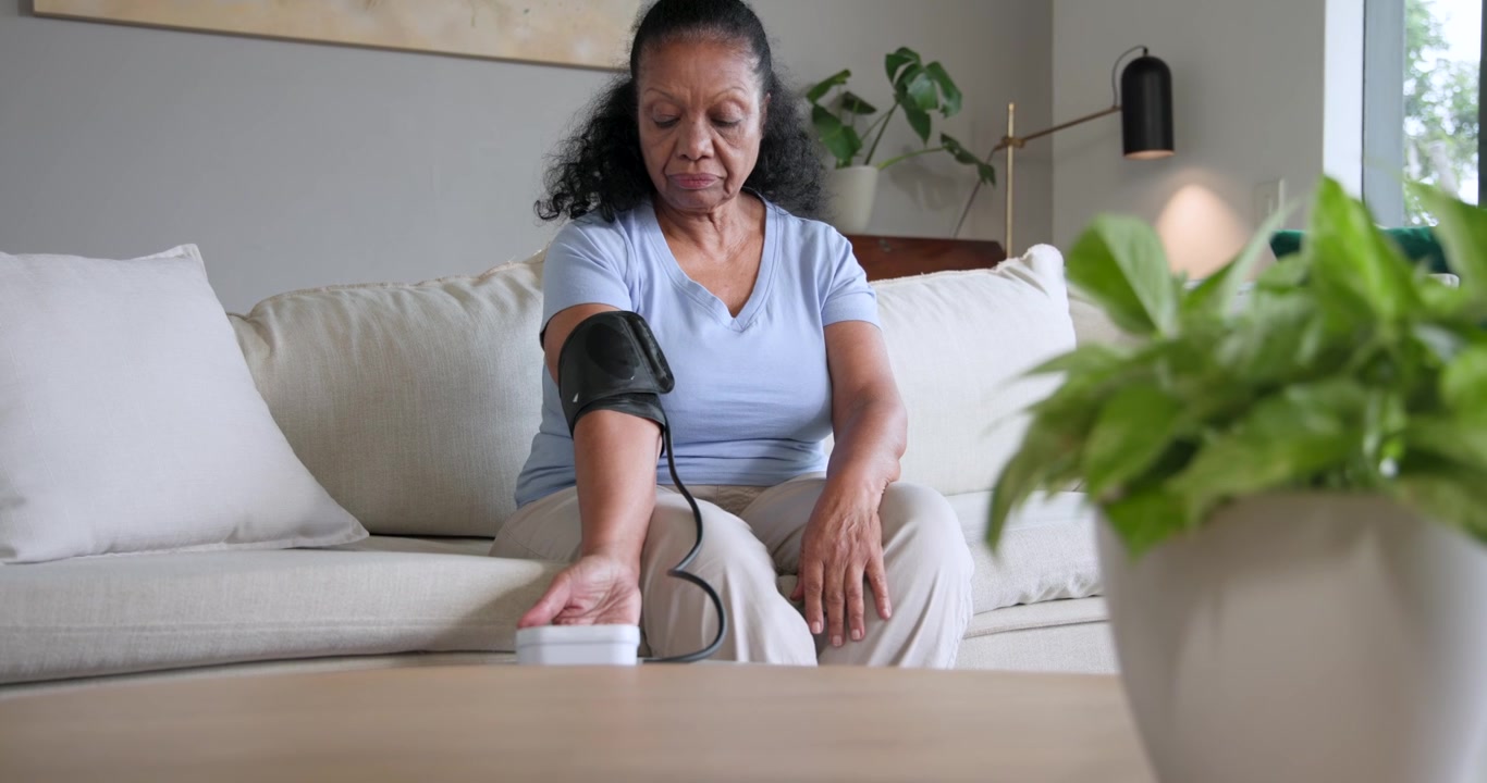 Donna afroamericana anziana che si prepara per un controllo della pressione sanguigna, posizionando il bracciale e usando un monitor bianco sul divano.