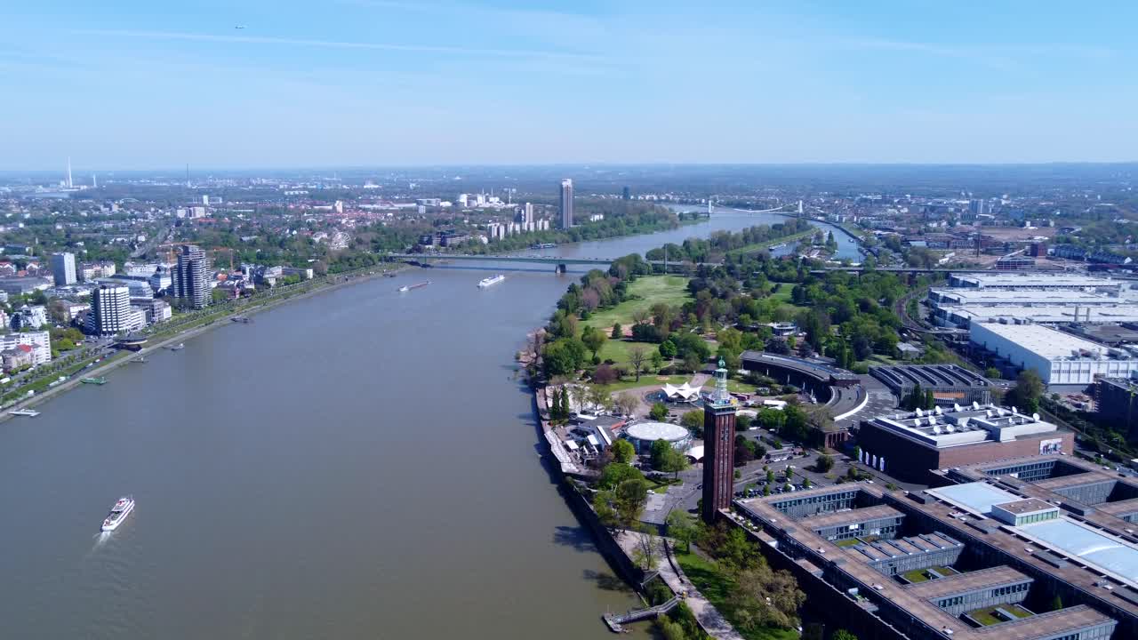 Veduta aerea con drone del lungofiume del Reno a Colonia con la Torre Rheinpark, ponti, aree verdi e barche. Chiaro paesaggio urbano diurno che mostra lo sviluppo urbano e l'infrastruttura del lungofiume.