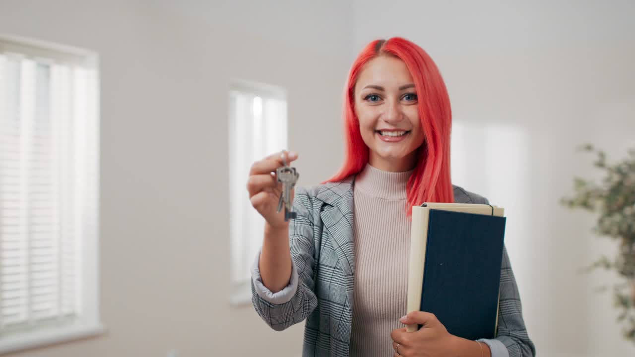Inquadratura di chiavi di appartamento tese su una mano verso la telecamera, donna dai capelli rosa vestita in tailleur tiene documenti e consegna le chiavi, agente immobiliare, proprietario dell'appartamento, consegna dell'immobile.