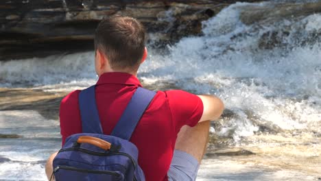 vista trasera de un viajero masculino en un polo rojo sentado junto a un río de montaña