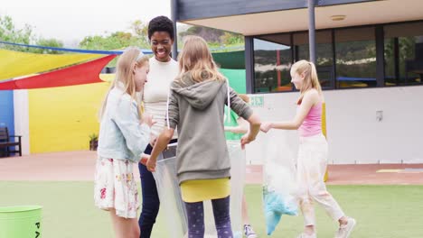 Diverse-female-teacher-and-happy-schoolchildren-cleaning-together-with-bags-at-school-playground