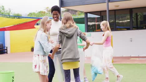 diverse female teacher and happy schoolchildren cleaning together with bags at school playground