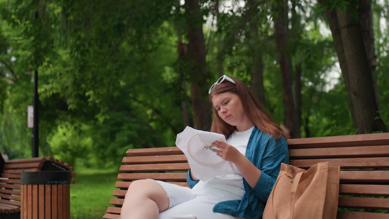 College girl sitting on wooden park bench carefully embroidering white fabric with needle, dressed in casual white outfit and blue shirt, focused on handmade craft in peaceful green park