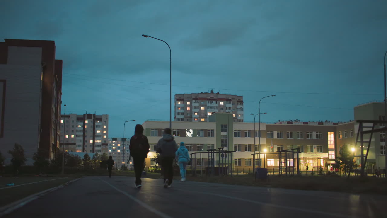 Children walking down school pathway in early morning before sunrise, backpacks on, surrounded by residential apartment buildings with windows lit, streetlights glowing, peaceful urban atmosphere