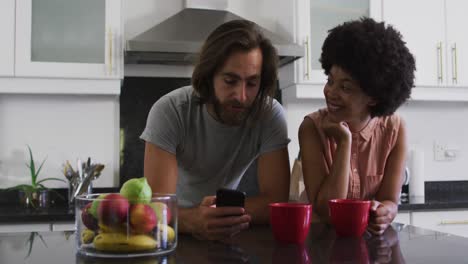 mixed race couple holding coffee cup using smartphone in the kitchen at home