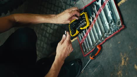 Vertical-video:-close-up:-a-male-mechanic-with-his-sinewy-hands-selects-the-tool-he-needs-among-a-set-of-tools-on-the-floor-in-the-workshop