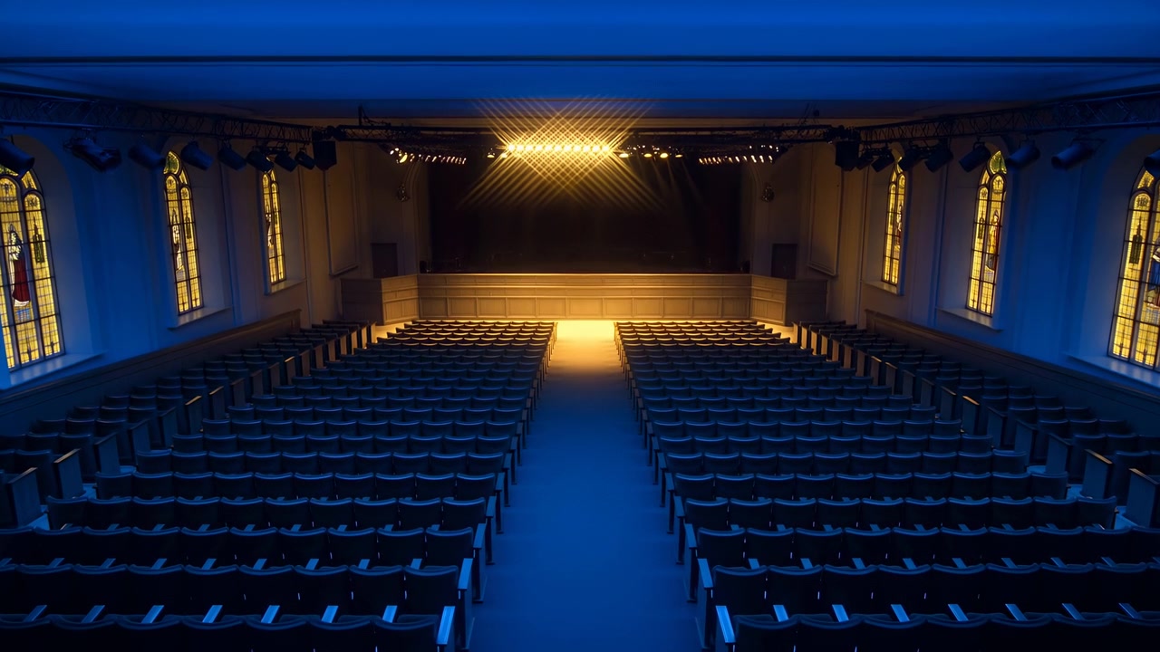 Un fotogramma intermedio mostra una vasta sala teatrale immersa in una luce blu. File vuote di sedili si estendono verso un palco luminoso, con un corridoio centrale che guida lo spettatore.