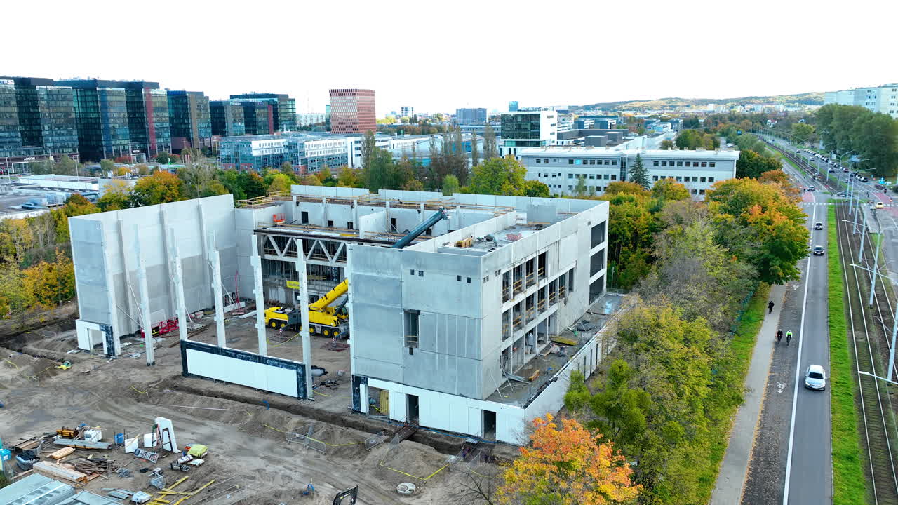 Aerial wide shot of a construction area and surrounding office buildings in Gdańsk, Poland, with modern architecture and a tree-lined street leading through the urban landscape