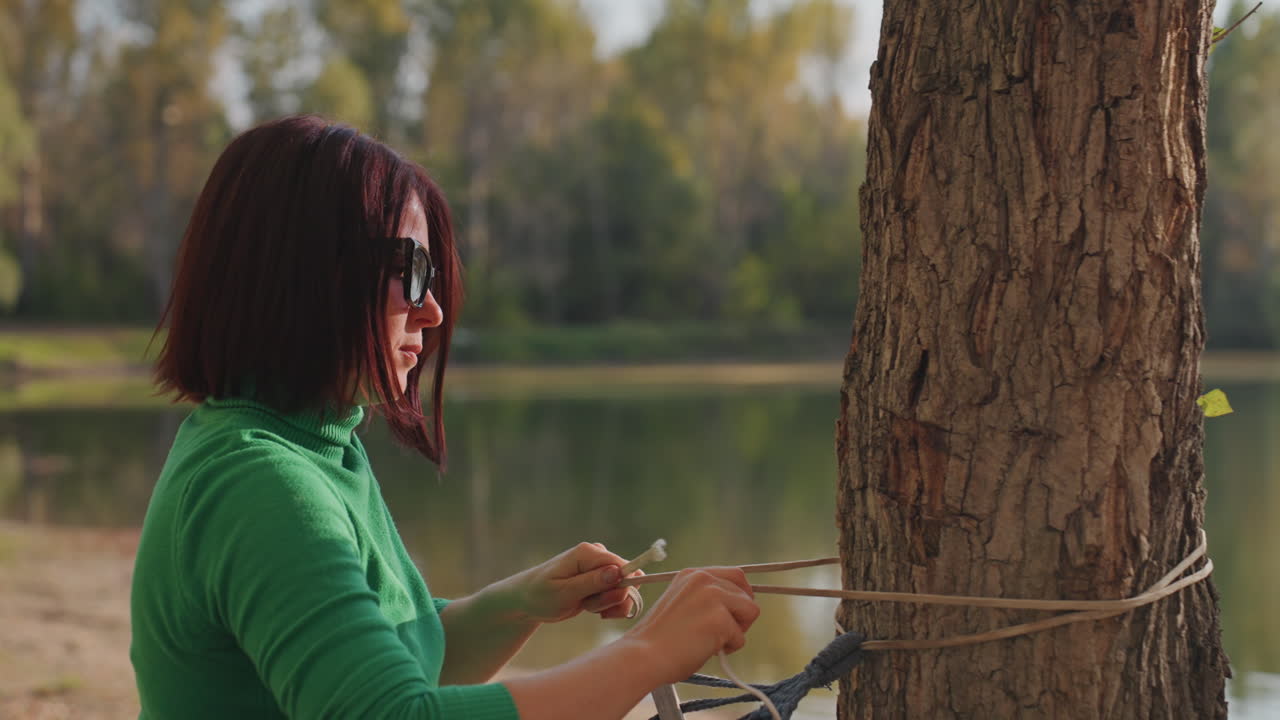 Woman sets hammock. Person prepares relaxing outdoor rest. Women stake out peaceful resting spot with careful knotting. Woman expertly secures hammock beside serene lakeside for leisure