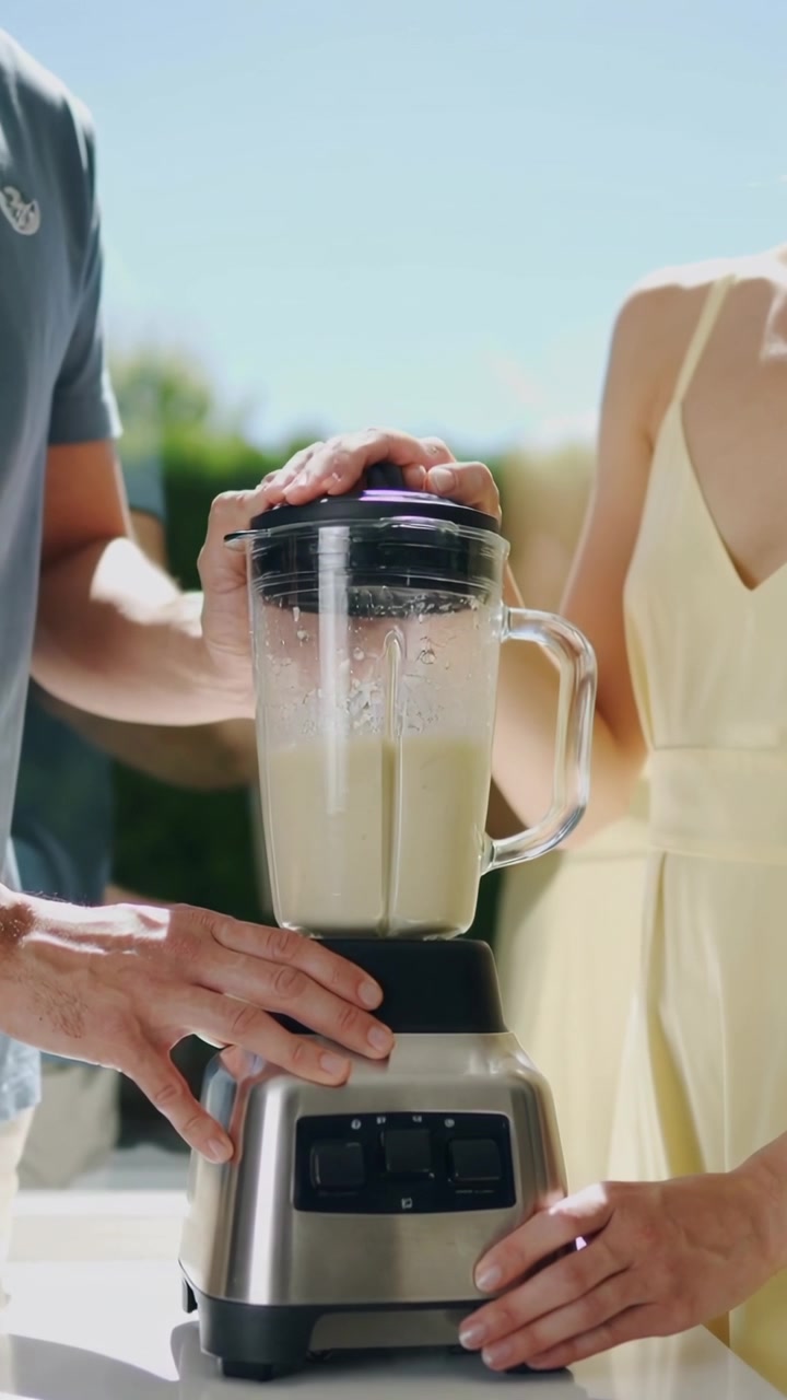 Uomo che aziona un comando e donna che tiene fermo il frullatore, mentre frullano un frullato verde per la preparazione in cucina all'aperto.
