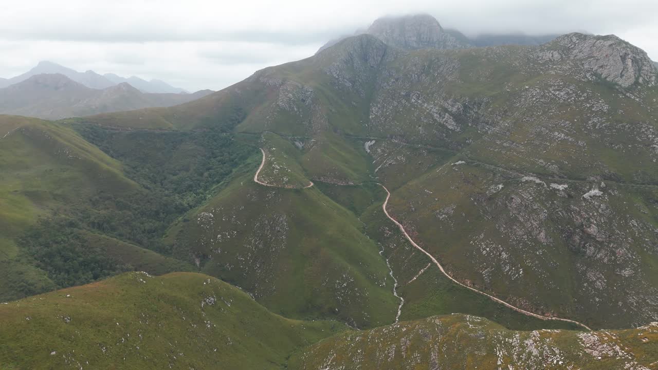 Aerial shot of the 7 Passes Hiking Trail along a mountain in Outeniqua Pass, Western Cape, South Africa. The drone follows the rugged terrain, revealing the scenic trail and surrounding natural beauty