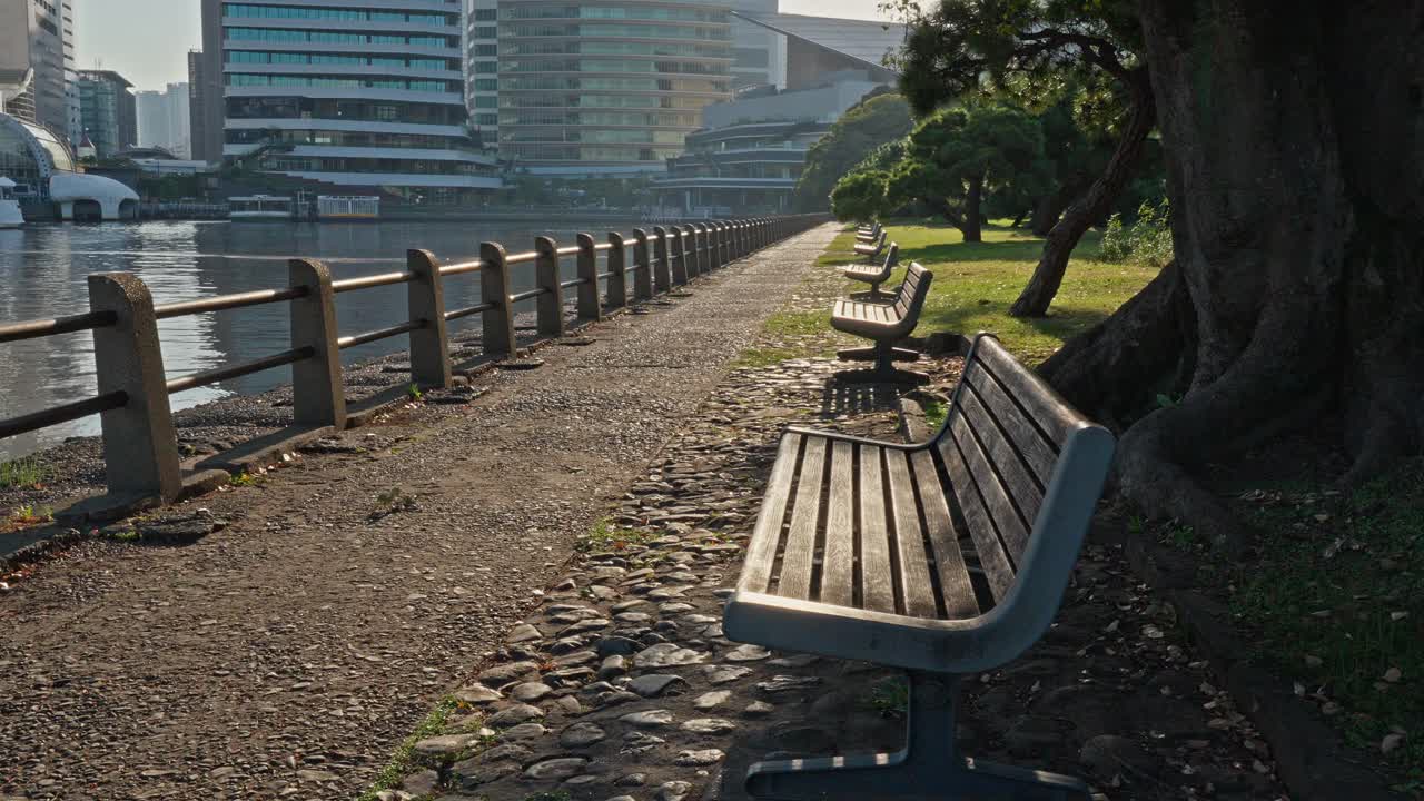 A long, paved path with benches lines a tranquil body of water in Hamarikyu Gardens, with modern city buildings in the distance.