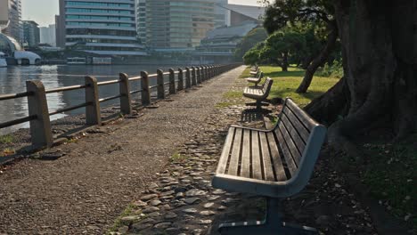 a long, paved path with benches lines a tranquil body of water in hamarikyu gardens, with modern city buildings in the distance.