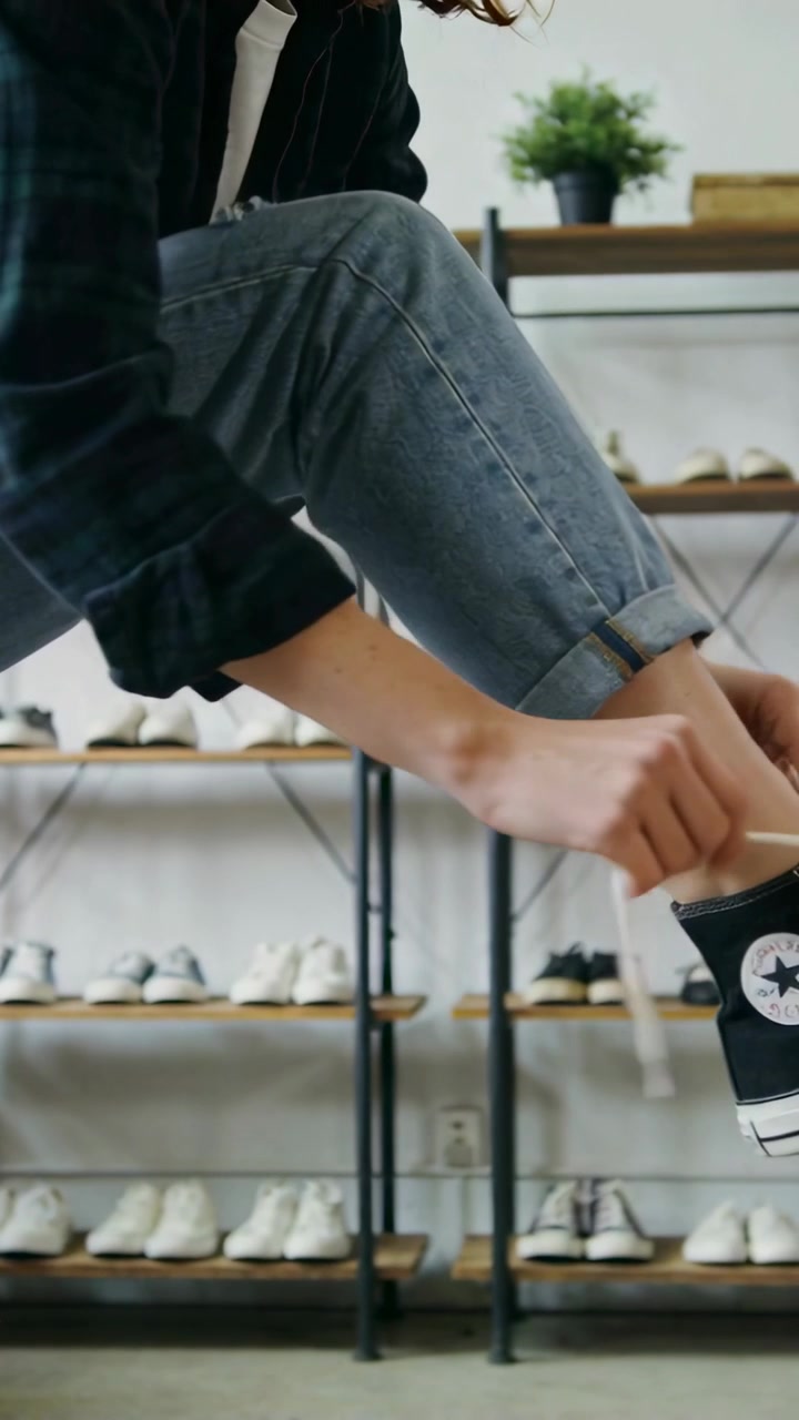 Vidéo verticale : Levant la jambe droite, un acheteur en jean nouant un lacet blanc sur une basket noire dans un magasin, prêt à marcher.