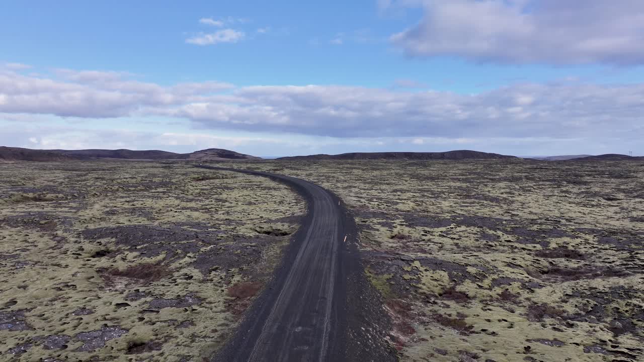 un pájaro ve un vehículo en una carretera sinuosa en terreno volcánico, bordeado por colinas y musgo, bajo un cielo sereno con nubes esponjosas