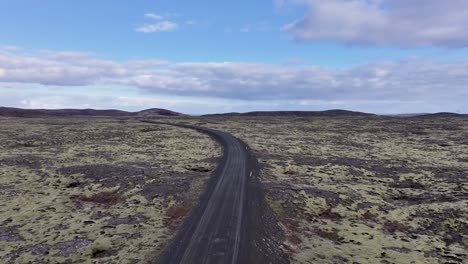 a bird sees a vehicle on a winding road in volcanic terrain, bordered by hills and moss, under a serene sky with fluffy clouds