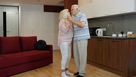 Feliz-Pareja-De-Ancianos-Maduros-Bailando-Riendo-En-La-Cocina-De-Casa,-Celebrando-El-Aniversario,-Divirtiéndose