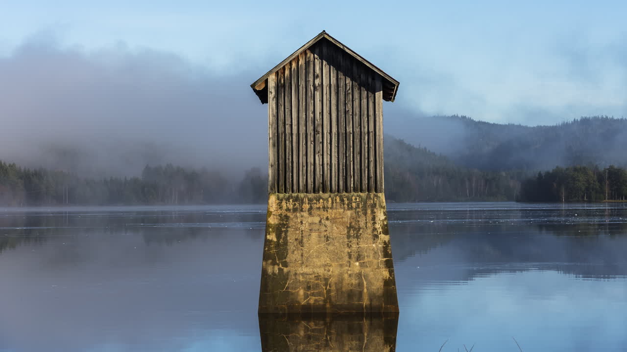 pequeña cabaña de madera antigua en medio de un lago tranquilo cubierto de niebla espesa
