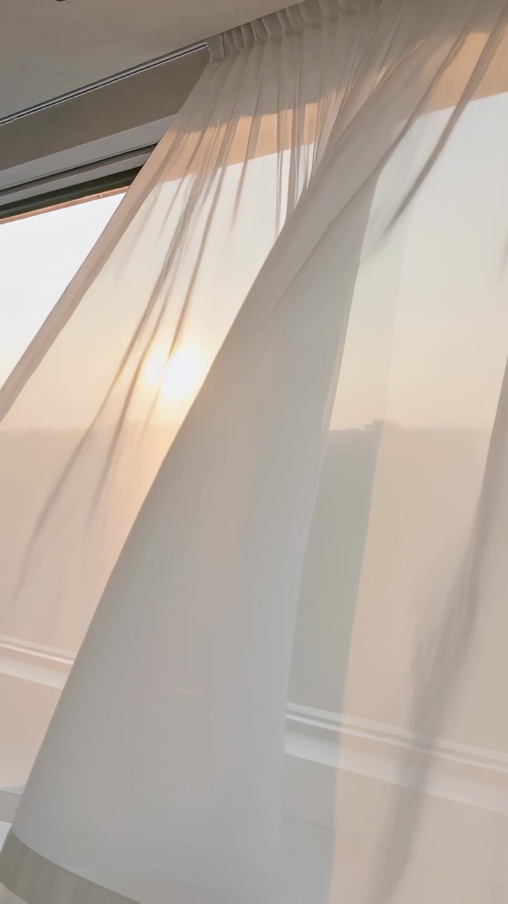 Vertical video: Breeze pushing sheer curtain billowing at home window, filtering warm sunset light