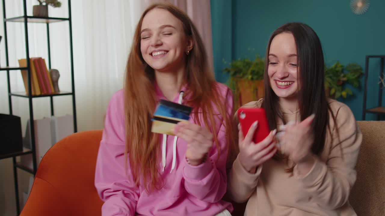 Cheerful girls friends using credit bank card and smartphone while transferring money purchases
