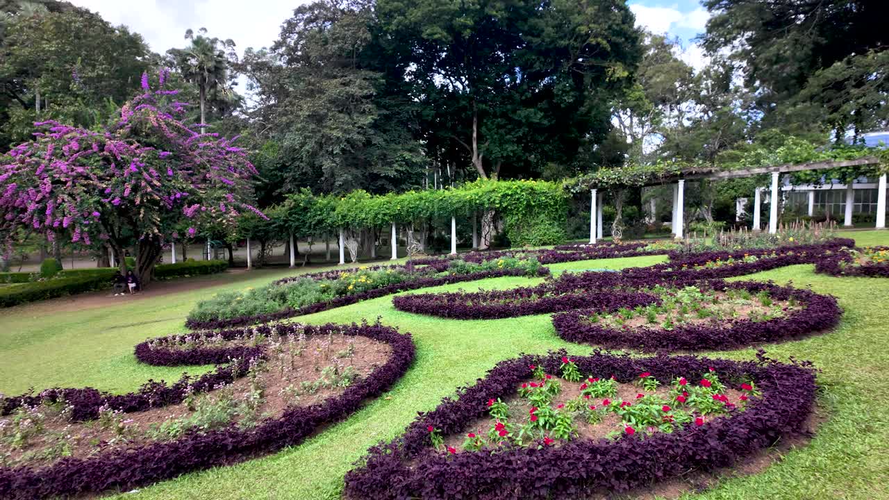 A tranquil garden with intricately shaped flower beds, lush green grass, and a backdrop of trees and pergolas, showcasing nature's beauty and serenity.