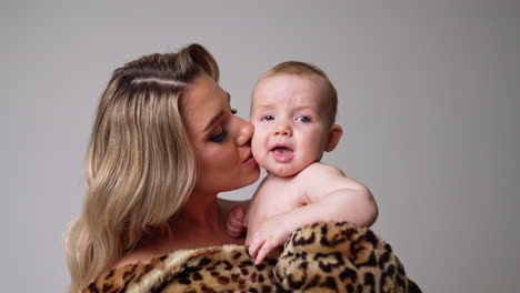 blonde caucasian blonde woman in fur coat holding a naked baby. child starting to cry and other kisses him to calm down the kid.