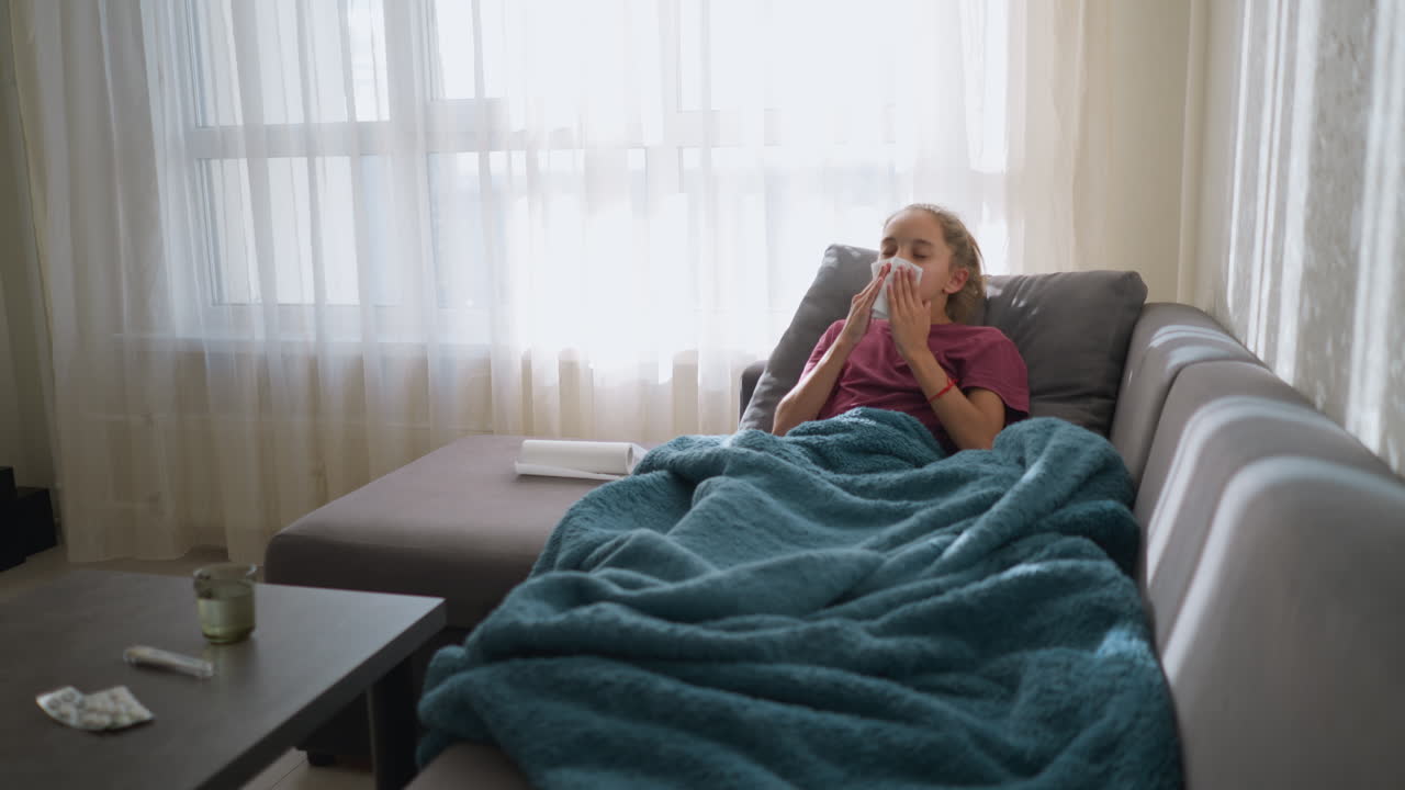 Female child in pink lying on couch suffering from flu, sneezing into tissue, with medication and glass cup on table, wrapped in blanket, looking unwell and tired