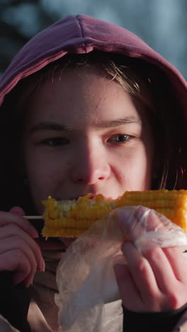 joven comiendo maíz en un palo envuelto en plástico blanco con luces bokeh en un fondo borroso, vapor que se eleva desde el maíz fresco, disfrutando de un bocadillo en un entorno al aire libre durante el invierno